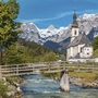 Kirche St. Sebastian in Ramsau vor Alpenkulisse und Holzbrücke über Fluss.