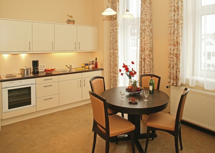 Küche im Haus Seeblick, in der Ferienanlage Ahlbeck, mit Esstisch, vier Stühlen, heller Küchenzeile mit Herd, Backofen und Spüle sowie einem Fenster und Balkontür mit Vorhängen.