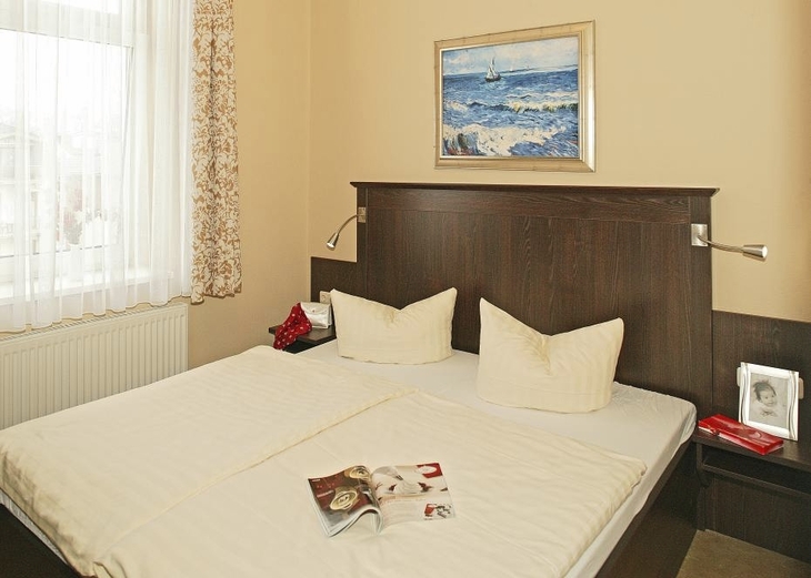 Schlafzimmer im Haus Seeblick, in der Ferienanlage Ahlbeck, mit Doppelbett, einem Bild darüber und einem Fenster mit weißer Gardine und gemustertem Vorhang.