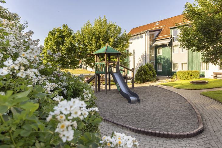 Spielplatz mit Rutsche in einer Gartenanlage vor einem Gebäude am sonnigen Tag.