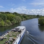 Flusskreuzfahrtschiff fährt auf einem von Bäumen gesäumten Fluss bei sonnigem Wetter.