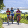 Drei Radfahrer auf einem Weg durch ländliche Landschaft mit See im Hintergrund.