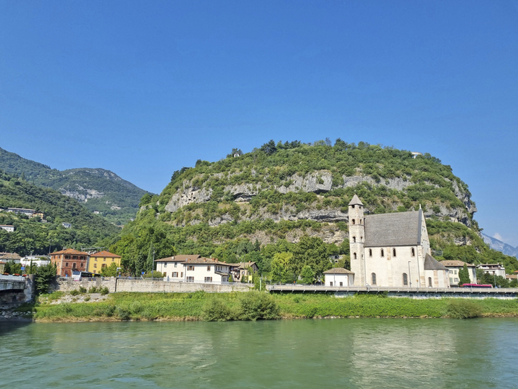 Kirche am Fluss vor bewaldetem Hügel und blauem Himmel.