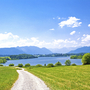 Weg führt durch grüne Wiesen zum Riegsee, Berge und blauer Himmel im Hintergrund.