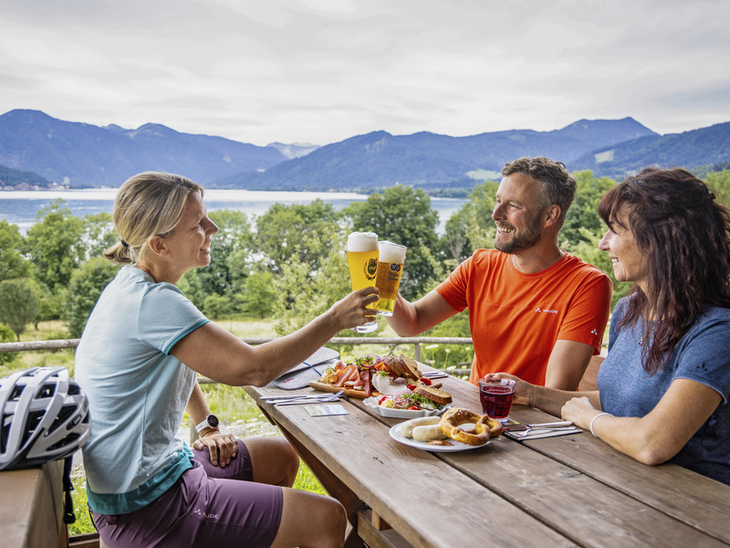 Drei Personen stoßen mit Bier an, im Hintergrund Tegernsee und Berge.