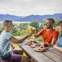 Drei Personen stoßen mit Bier an, im Hintergrund Tegernsee und Berge.