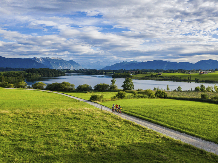 Radfahrer auf einem Weg neben dem Riegsee, umgeben von grüner Landschaft und Bergen.