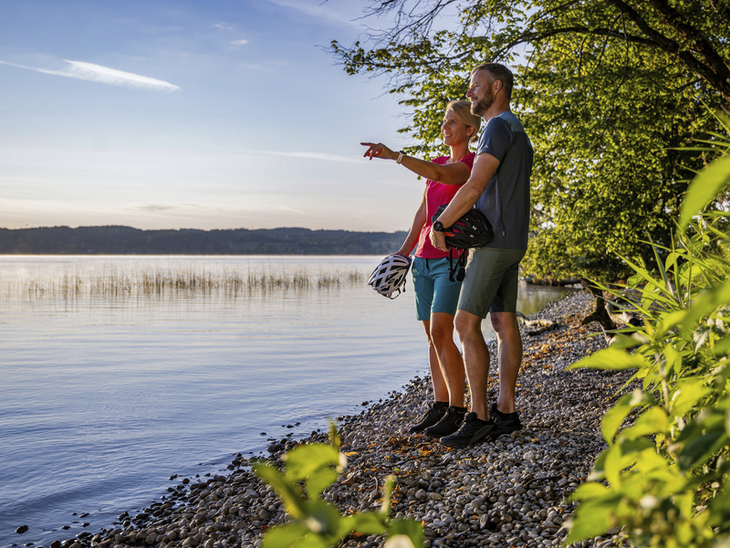 Paar macht Radpause am Starnberger Seeufer, schaut auf das Wasser bei sonnigem Wetter.