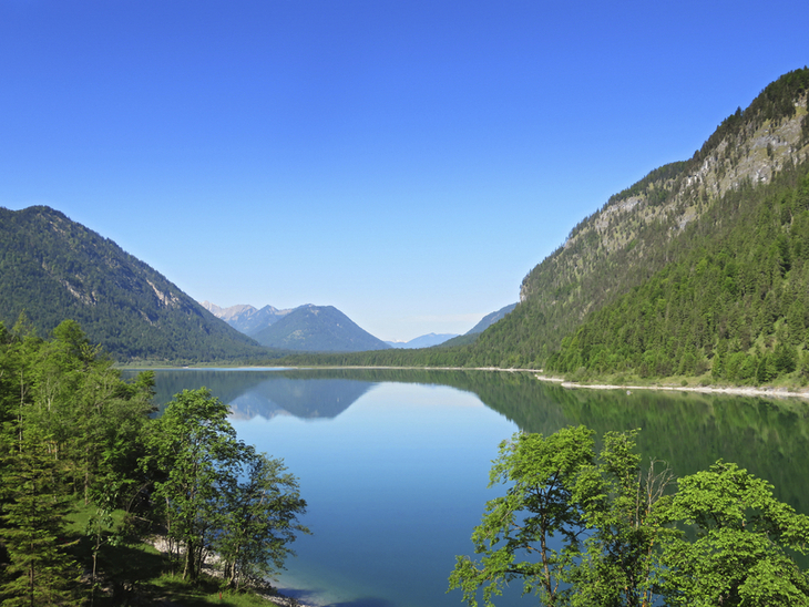 Blick auf den klaren Sylvensteinspeicher mit umliegenden bewaldeten Bergen bei blauem Himmel.