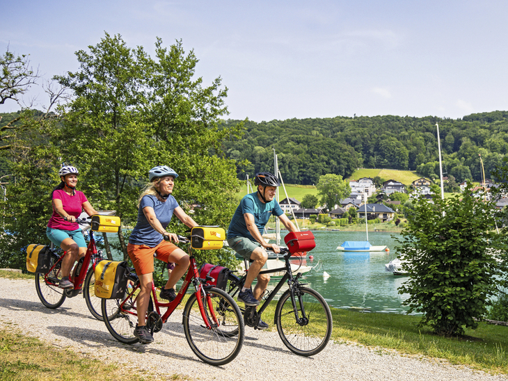 Drei Radfahrer fahren auf einem Weg entlang eines Sees mit im Hintergrund sichtbaren Booten.