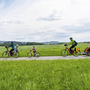 Familie radelt auf einem Weg durch eine grüne Landschaft, Kinderanhänger dabei.