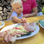 Kind greift nach Speckbrot auf Holztisch im Freien.