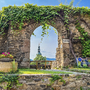 Blick durch ein bewachsenes Steinportal auf einen Kirchturm in Krems, umgeben von Pflanzen.