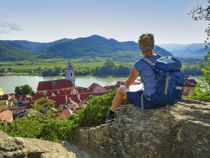 Wanderin betrachtet Donaublick von Ruine Dürnstein, umgeben von Hügeln und Flusslandschaft.