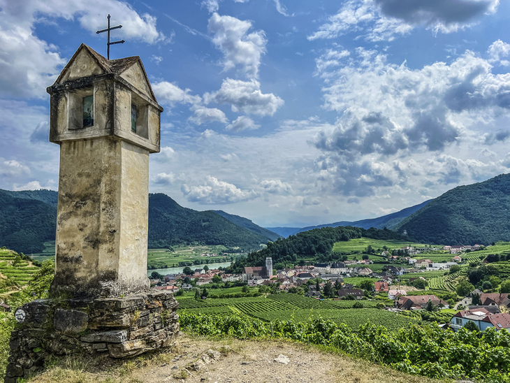 Kleines Denkmal am Hang, Blick auf grünes Tal mit Dorf und Bergen im Hintergrund.