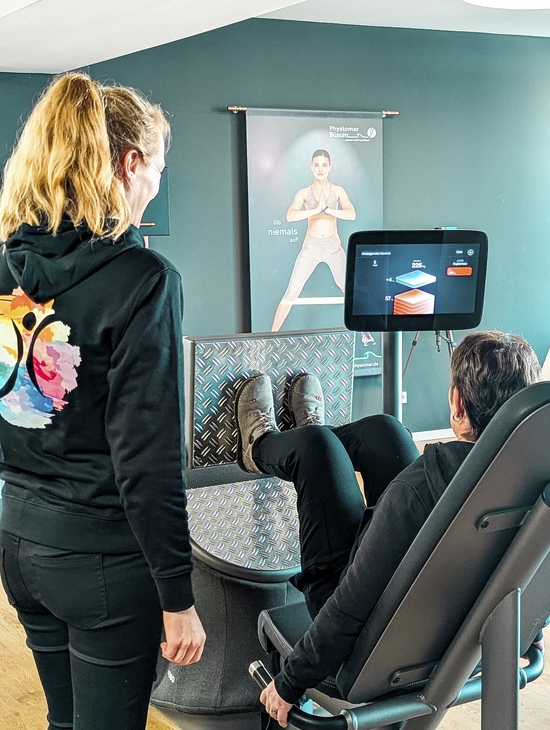 Eine Person trainiert an einem Fitnessgerät, betreut von einer Trainerin.