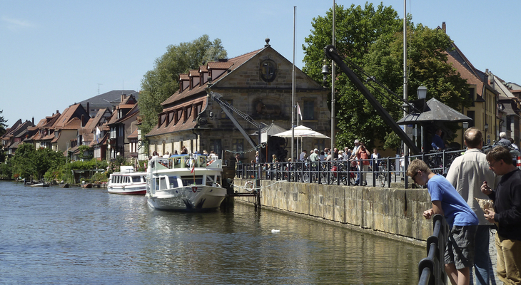 Menschen und Boote entlang einer Promenade am Fluss in Bamberg bei sonnigem Wetter.