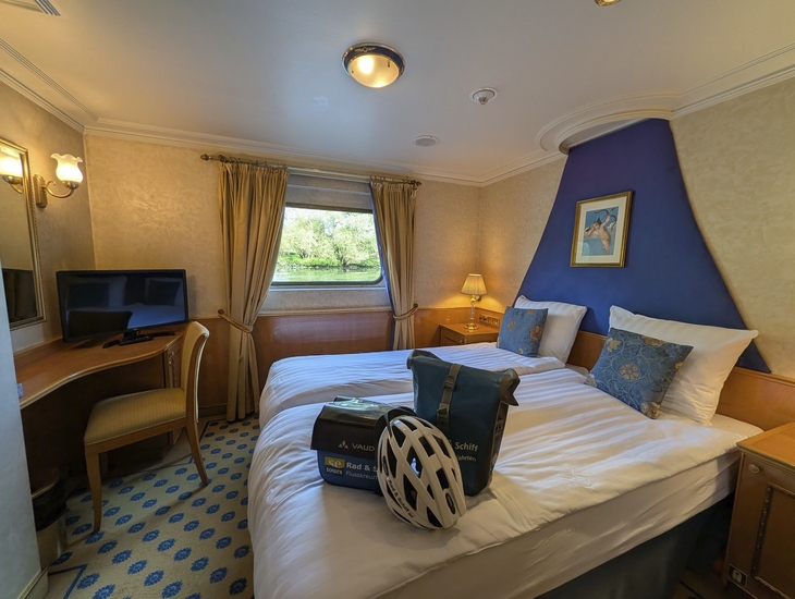 Kabine mit Doppelbett, Schreibtisch, Fenster und Fahrradhelm auf dem Bett.